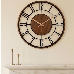 May include: A round wall clock featuring a dark brown wooden center disc and a black metal frame with large, open-style Arabic numerals. The clock has two slender, metallic gold-toned hands positioned at the center, indicating the time.