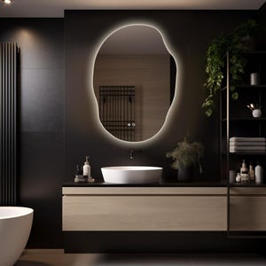 May include: An irregular, organic-shaped wall mirror featuring a built-in LED backlight that creates a soft glow around its perimeter. The mirror is mounted above a white vessel sink and a light wood vanity cabinet in a dark-toned bathroom.