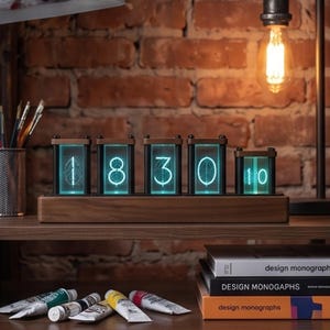 Könnte beinhalten: Eine Digitaluhr mit fünf vertikalen Glasanzeigefeldern auf einem rechteckigen Holzsockel. Die Paneele leuchten in einem hellen Cyan und zeigen die Ziffern 1, 8, 3, 0 und 1 mit sichtbaren, komplexen geometrischen Linienmustern im Inneren.