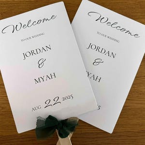 May include: Two white wedding programs with the words "Welcome" and "Jordan & Myah" printed in black script. The date "Aug 22 2025" is also printed on the programs. A dark green ribbon is attached to the bottom.