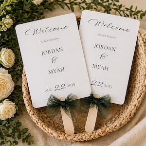 以下が含まれることがあります： 「Welcome」の文字と「Jordan & Myah」の名前、日付「2025年8月22日」が書かれた白いウェディングファン2つ。各ファンには濃い緑色のリボンと木製の持ち手が付いています。ファンは編まれたバスケットに入っています。