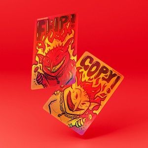 May include: Two rectangular game tokens with rounded corners, featuring a red and orange color scheme. One token displays the text FLIP! above a grinning creature with a chain, while the other shows the text COPY! above a similar creature.