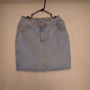 May include: A light blue denim skirt with a button fly and two pockets on the front. The skirt is hanging on a black hanger against a white wall.