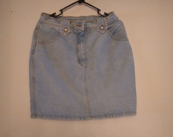 May include: A light blue denim skirt with a button fly and two pockets on the front. The skirt is hanging on a black hanger against a white wall.