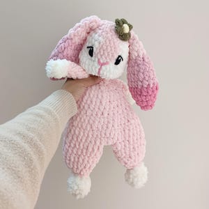 May include: A handmade pink and white crochet bunny plush toy with a flower accent. The bunny has long ears, black eyes, and white pom-poms on its feet. The plush is held up by a person's hand.