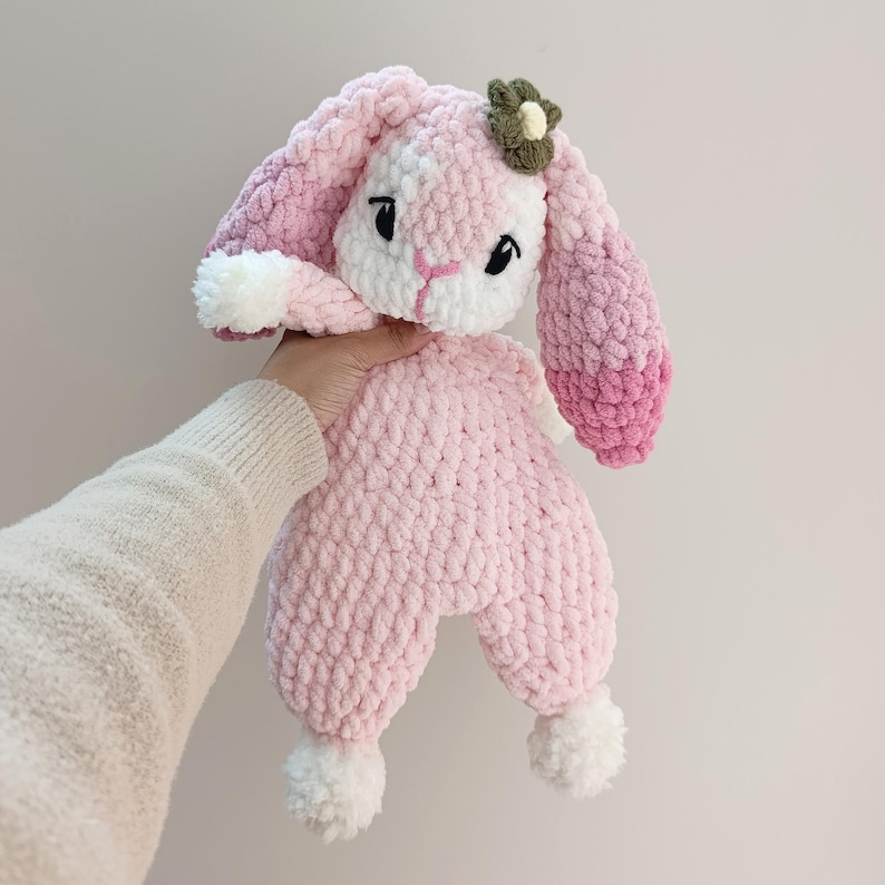 May include: A handmade pink and white crochet bunny plush toy with a flower accent. The bunny has long ears, black eyes, and white pom-poms on its feet. The plush is held up by a person's hand.