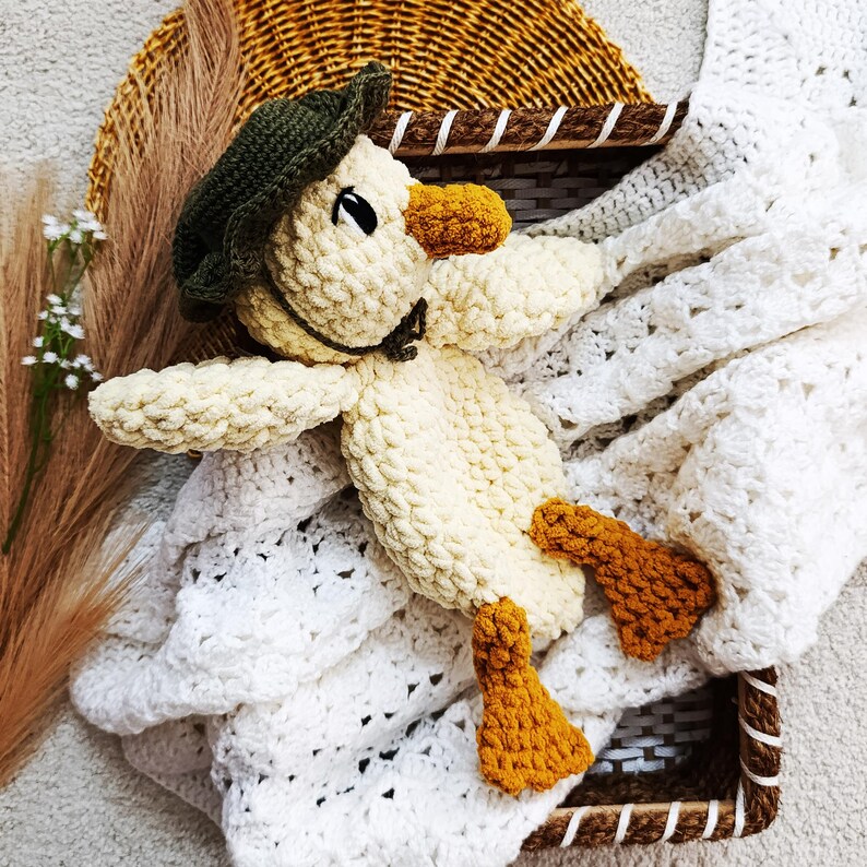 May include: A handmade crochet duck toy in cream, with an orange beak and feet, and a green cap. The duck is lying on a white crocheted blanket inside a woven basket. The background includes dried plants.