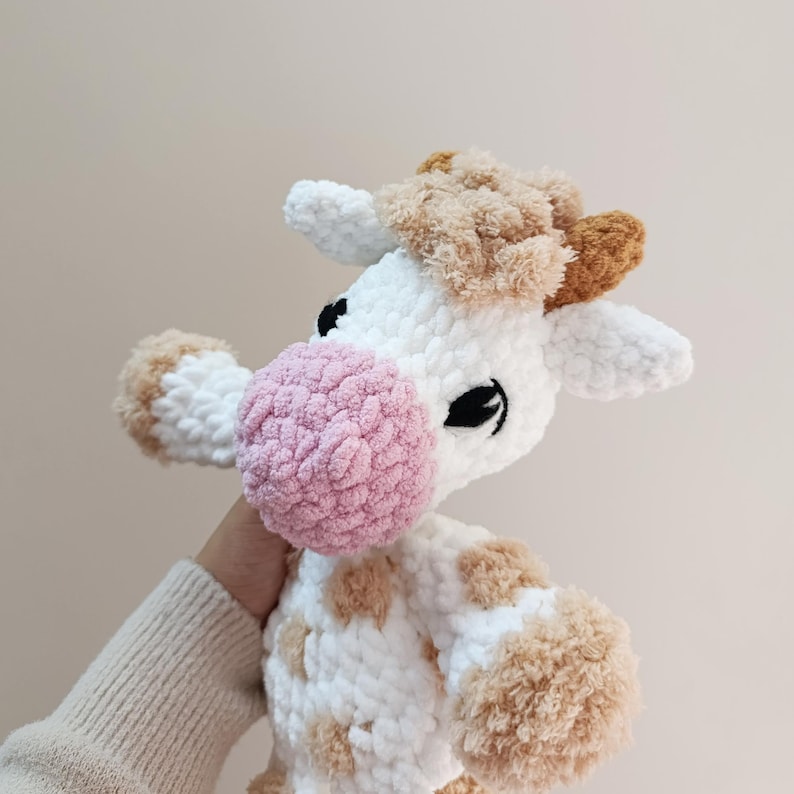 May include: A handmade crochet cow plushie. The cow is white with brown spots and accents on the ears and head. It has a pink nose and black eyes. The plushie is soft and fluffy, perfect for a child's toy or nursery decor.
