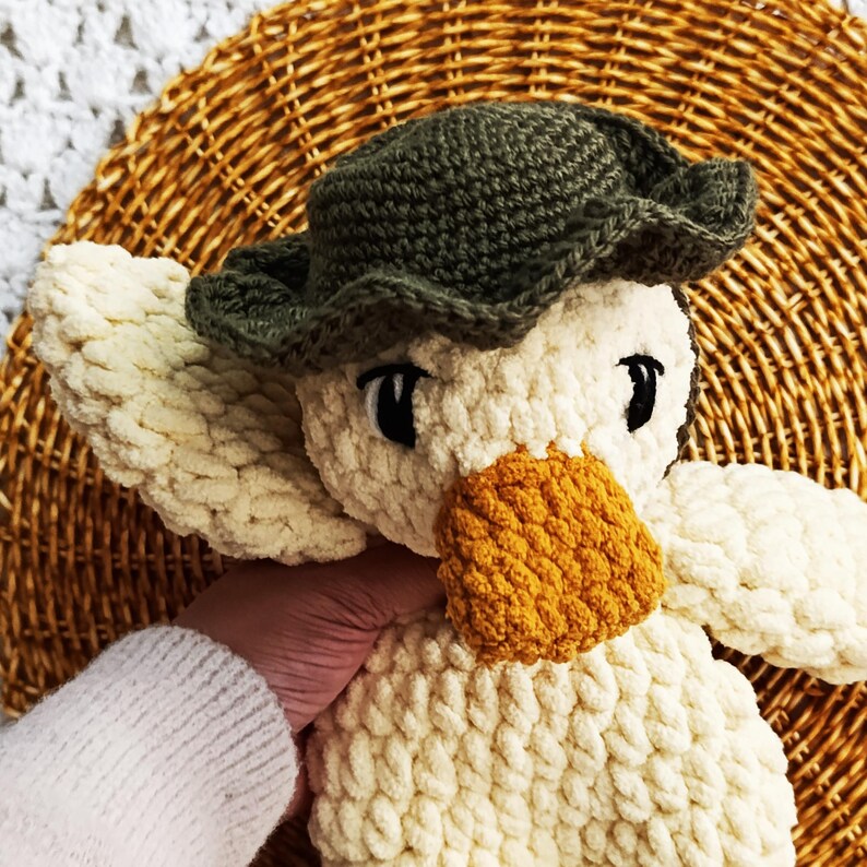 May include: A handmade, plush duck toy with a green hat and an orange beak. The duck is made of cream-colored yarn and has black eyes. The toy is designed to be soft and cuddly.