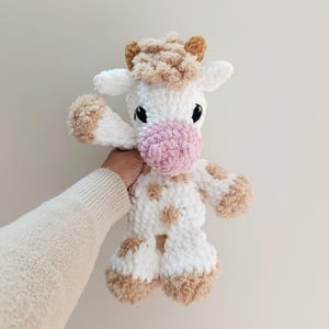 May include: A handmade plush cow toy with a white body, brown spots, and a pink nose. The cow has black eyes and brown horns. The toy is made of soft, textured yarn and is designed to be cuddly.