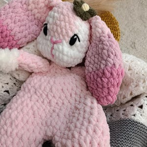 May include: A handmade, plush bunny in shades of pink and white. The bunny has long, floppy ears with a gradient of pink, black eyes, and a small flower on its head. The soft toy is made of a textured yarn.