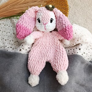 May include: A handmade pink crochet bunny toy with long, ombre pink ears and white accents. The bunny has black eyes and a small green and brown flower on its head. The toy is lying on a gray and white crocheted blanket.