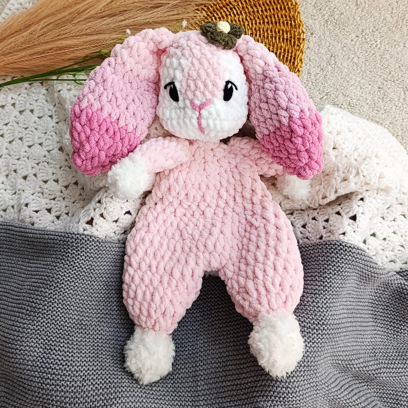 May include: A handmade pink crochet bunny toy with long, ombre pink ears and white accents. The bunny has black eyes and a small green and brown flower on its head. The toy is lying on a gray and white crocheted blanket.