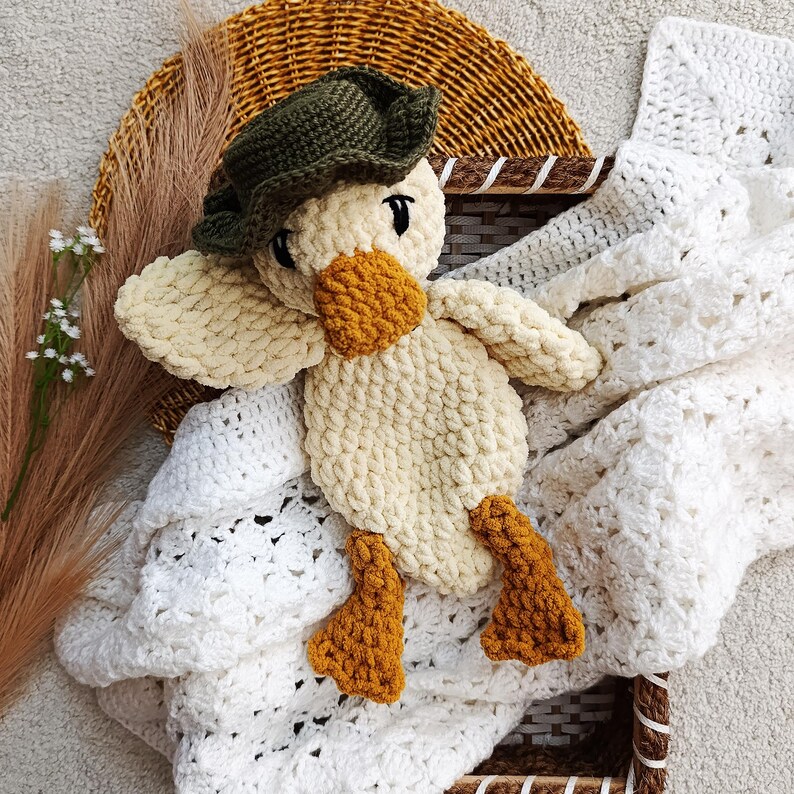 May include: A handmade crochet duck toy with a cream-colored body, orange beak and feet, and a green hat. The duck is lying on a white crocheted blanket inside a wicker basket. The toy is soft and plush.