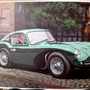 May include: A vintage green sports car with a dark green stripe along the side. The car has wire wheels and is parked on a street with buildings and a sign that reads "Keep Left". A person is visible inside the car.