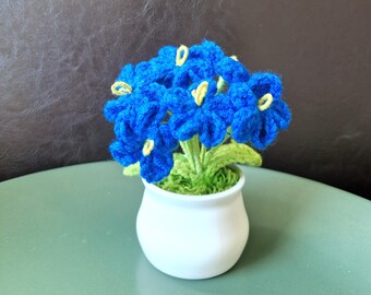 May include: A handmade crochet flower arrangement featuring vibrant blue flowers with yellow centers and green leaves. The flowers are in a small white ceramic pot, creating a charming decorative piece.