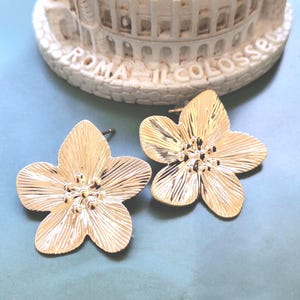 May include: A pair of gold-colored flower-shaped stud earrings. The earrings have detailed petal designs and a textured finish. The background includes a white decorative piece with the text "ROMA IL COLOSSEO".
