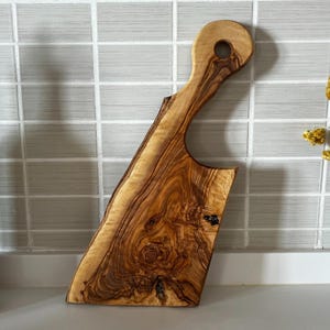 Op de afbeelding: Een houten snijplank met een natuurlijke rand en een handvat met een gat. De plank is gemaakt van olijfhout en heeft een rijke, warme bruine kleur met unieke nerfpatronen. De plank is ongeveer 38 cm lang.