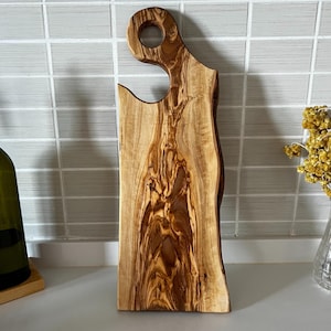 May include: A wooden cutting board with a natural edge and a handle with a hole. The board is made of olive wood, displaying a rich, brown grain pattern. The board is approximately 12 inches long and 6 inches wide.
