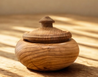 Peut inclure: Un petit récipient rond en bois avec un couvercle. Le récipient et le couvercle sont en bois brun clair, avec le grain du bois visible. Le couvercle a un petit bouton décoratif sur le dessus. Le récipient repose sur une surface en bois.