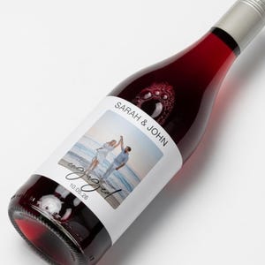 May include: A glass wine bottle filled with deep red liquid, featuring a custom white label with a photograph of a couple on a beach, the text SARAH & JOHN, the word engaged in script, and the date 10.05.26, topped with a silver screw cap.
