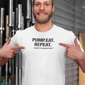 Könnte beinhalten: Ein weißes T-Shirt mit dem schwarzen Text "PUMP.EAT. REPEAT. EVERY FU##N DAY!". Die Person zeigt auf das T-Shirt. Der Hintergrund umfasst Gewichtheberausrüstung.