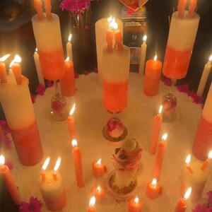 May include: An arrangement of lit candles in varying sizes and shapes, including tall, two-toned pillar candles and slender orange tapers. The candles are arranged on a white surface, surrounded by pink flowers and a heart-shaped picture.