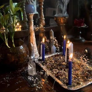 May include: A mystical scene featuring a silver tray with burning candles, dried herbs, and flowers. Tall, ornate candle holders and a potted plant are in the background. A blue orb sits atop a decorative stand, and a tarot card is visible.