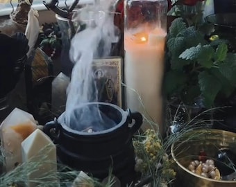 May include: A black cast iron cauldron emitting white smoke, positioned next to a tall cylindrical glass candle with a glowing flame. The scene includes several light-colored crystal points, a small gold bowl with pearls, and green foliage.
