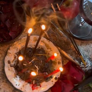 May include: A still-life arrangement featuring a lit candle display on a decorative plate, surrounded by red roses, apples, and a glass of red wine. Smoke rises from the candles, creating a mystical ambiance. Incense sticks and a bowl of petals are also present.