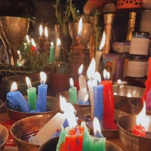 May include: A collection of lit candles in various colors, including blue, green, red, and white, are arranged in small, round, brass-colored bowls. The candles are of varying heights and are placed on a table, creating a warm, inviting atmosphere.