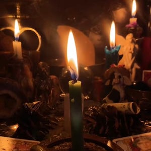 May include: A green taper candle burns with a bright flame in the foreground. Surrounding the candle are various objects including a small cow figurine, a rolled-up dollar bill, pinecones, and tarot cards under dim, warm lighting.