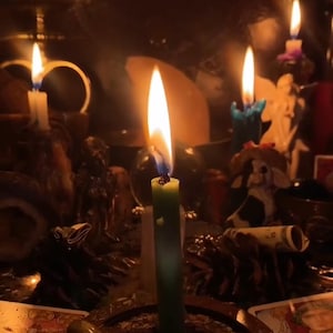 May include: A dimly lit scene featuring several lit candles of varying colors and sizes. A green candle is in focus, with a bright flame. Other candles are visible, along with decorative items, creating a mystical ambiance.