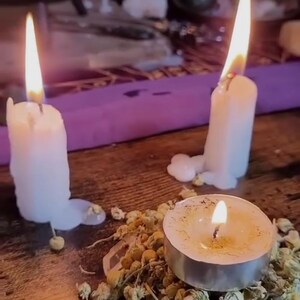 May include: Three lit candles of varying sizes on a wooden surface. Two are tall, white, and dripping wax. A small, round tealight candle is also lit, surrounded by dried herbs. A purple cloth is in the background.