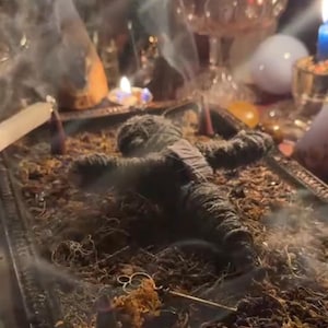 May include: A dark gray voodoo doll lies on a bed of dried moss and herbs within a decorative tray. Smoke rises from the scene, with lit candles and other ritualistic items in the background, creating an atmospheric setting.