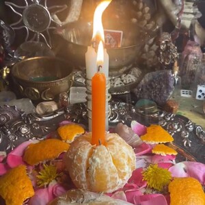 May include: An orange candle is lit, placed atop a peeled tangerine, surrounded by pink rose petals and orange peel. The scene includes a silver tray, crystals, and other ritualistic items, creating a mystical ambiance.
