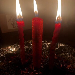 May include: Three red candles with bright flames burning in a dimly lit setting. Two candles have a twisted design, while the center candle is straight. The candles are placed on a foil-lined surface.