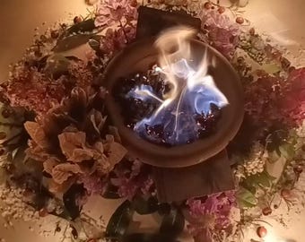 May include: A dark brown ceramic bowl containing a bright blue flame, surrounded by a circular arrangement of dried pink and brown flowers, green leaves, small brown berries, and white salt, illuminated by four small white candles.