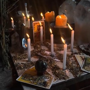 May include: A mystical arrangement with lit candles, tarot cards, and a crystal ball. White and orange candles are placed on a surface with dried herbs. Tarot cards with illustrations are scattered around, creating a mysterious atmosphere.