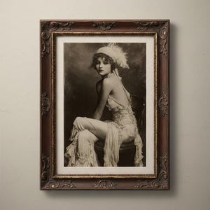 May include: A framed black and white photograph of a woman in a vintage dress and feathered headpiece. The dark brown frame has ornate detailing. The woman is seated on a chair, with a soft, blurred background.