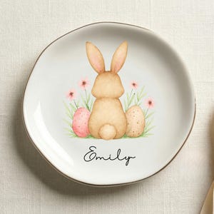 May include: A white ceramic plate with a gold rim featuring a watercolor illustration of an Easter bunny, two eggs, and pink flowers. The name "Emily" is written in cursive below the illustration. The plate is on a cream-colored surface.