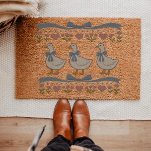 May include: A rectangular doormat made of natural coir fiber. The mat features three cartoon geese wearing blue bows, with a decorative border of hearts, flowers, and ribbons. The mat is a light brown color.