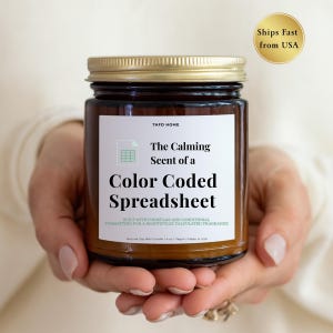 May include: A brown glass jar candle with a gold lid, held in hands. The label reads "The Calming Scent of a Color Coded Spreadsheet." The candle is made with natural soy wax and is vegan. The text "TAFD HOME" is at the top of the label.