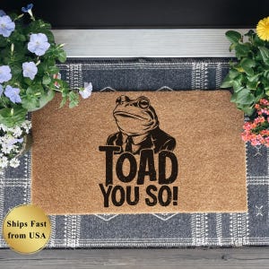 May include: A rectangular coir doormat with a black graphic of a toad in a suit and tie. The text "TOAD YOU SO!" is printed below the toad. The mat is surrounded by potted blue and yellow flowers, and rests on a patterned rug.