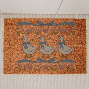 May include: A rectangular doormat with a textured orange background. Three gray geese with blue bows are centered on the mat. Above and below the geese are blue ribbons, purple hearts, and green floral accents. The mat is bordered by a white fabric.