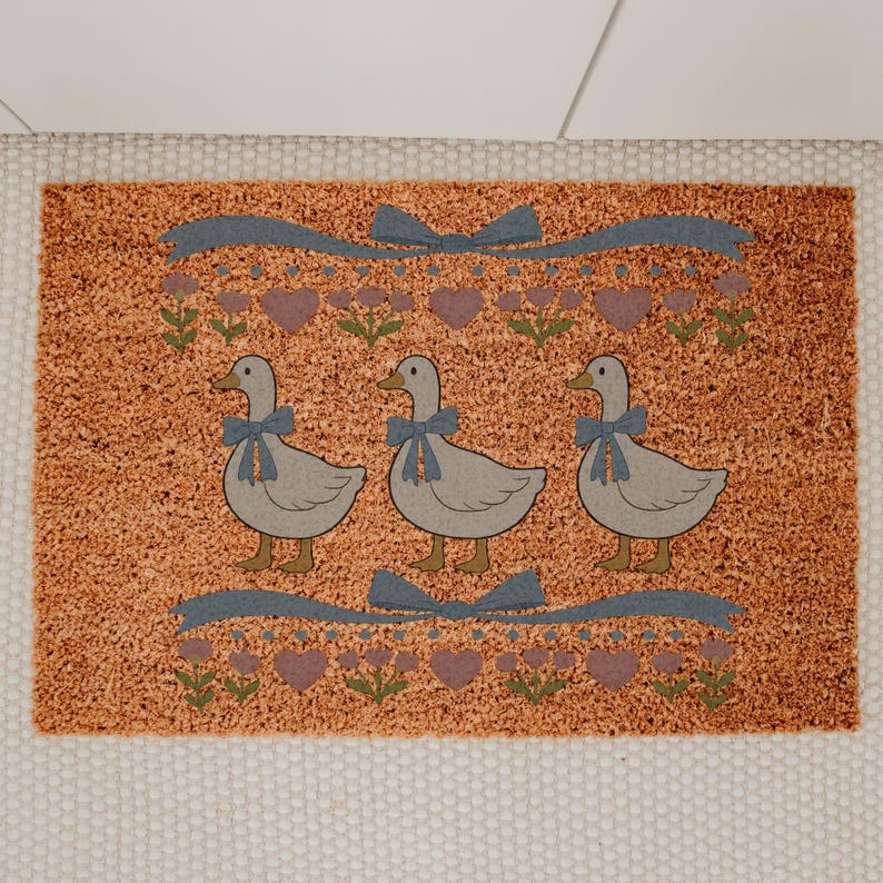 May include: A rectangular doormat with a textured orange background. Three gray geese with blue bows are centered on the mat. Above and below the geese are blue ribbons, purple hearts, and green floral accents. The mat is bordered by a white fabric.