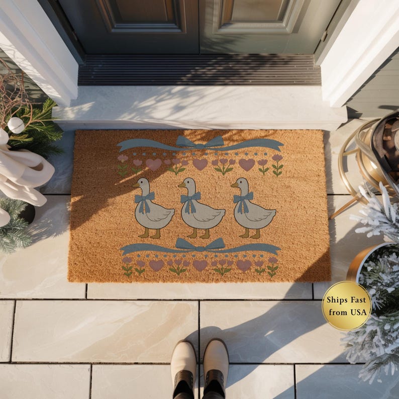 May include: A rectangular doormat with a light brown background. The mat features three cartoon geese with blue bows, surrounded by hearts, flowers, and ribbons. The mat is placed in front of a dark gray door.