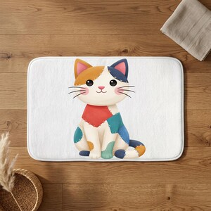 May include: A rectangular bath mat featuring a white background with a central illustration of a sitting cat. The cat is designed with a patchwork appearance, displaying various colors including orange, blue, teal, and red, with visible stitching.
