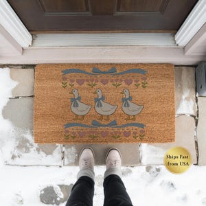 May include: A rectangular doormat with a light brown background features three cartoon geese wearing blue bows. The design includes blue ribbons, pink hearts, and floral accents. The mat is placed in front of a brown door on a stone surface.