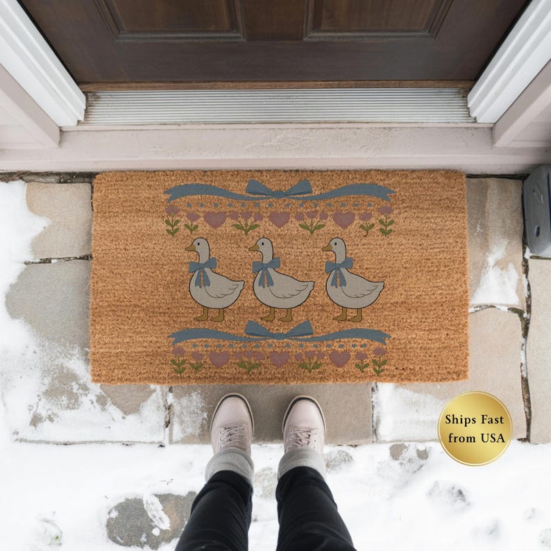 May include: A rectangular doormat with a light brown background features three cartoon geese wearing blue bows. The design includes blue ribbons, pink hearts, and floral accents. The mat is placed in front of a brown door on a stone surface.